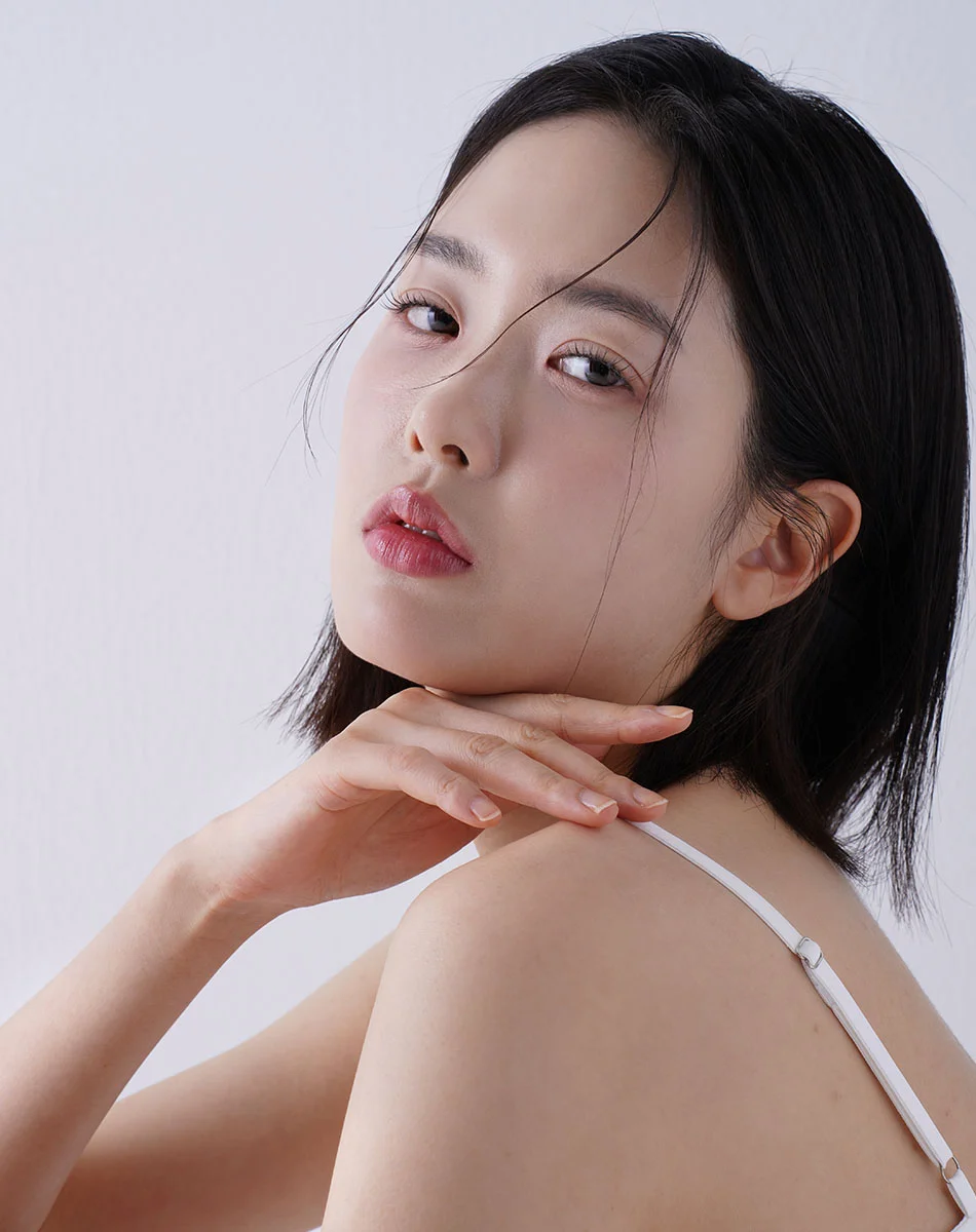 Close-up of a young woman with short dark hair, neutral expression, and light-toned skin, gently resting her hands on her neck and shoulder, wearing a white strap top against a light gray background. - Acne Scar Treatment in Washington, DC and New York, NY