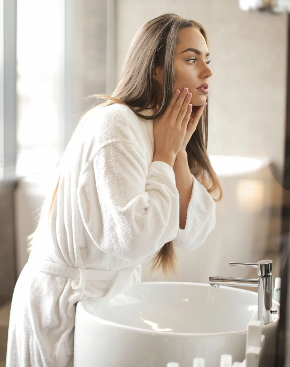 Young woman in a white bathrobe stands in a bright bathroom, examining her face in the mirror with hands near her cheeks. - Aviclear Laser Treatment for Acne in Washington, DC and New York, NY
