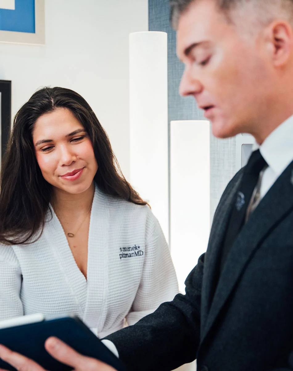 Woman in a white bathrobe with “Physician MD” embroidered on it smiles while holding a tablet, seated next to a man in a dark suit with his arm around her; they are in a medical or spa office setting. - Breast Augmentation in Washington, DC and New York, NY