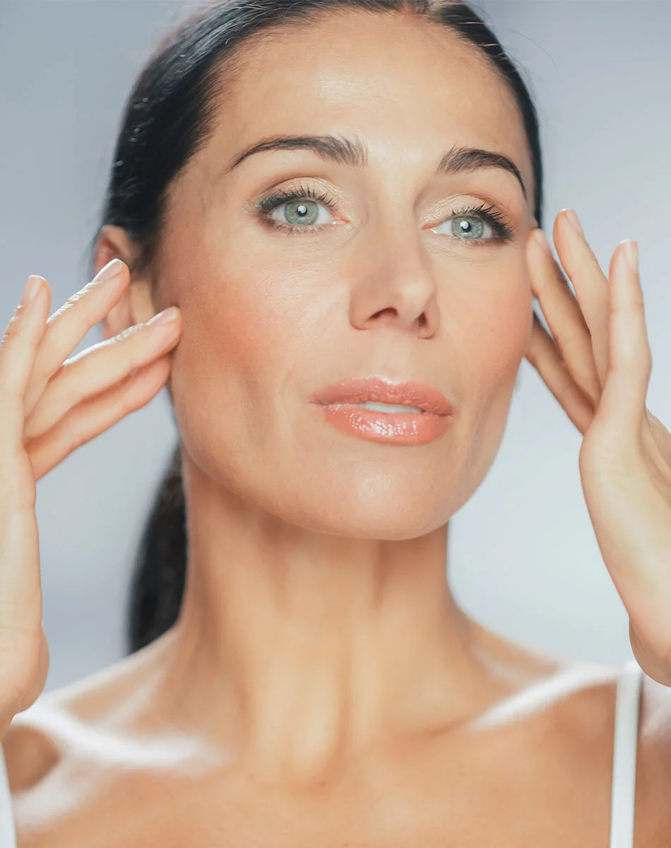 Close-up of a light-skinned woman with dark hair pulled back, gently touching her cheeks. She wears light makeup and gazes neutrally at the camera. Soft lighting highlights her smooth skin against a muted gray background. - Buccal Fat Removal in Washington, DC and New York, NY