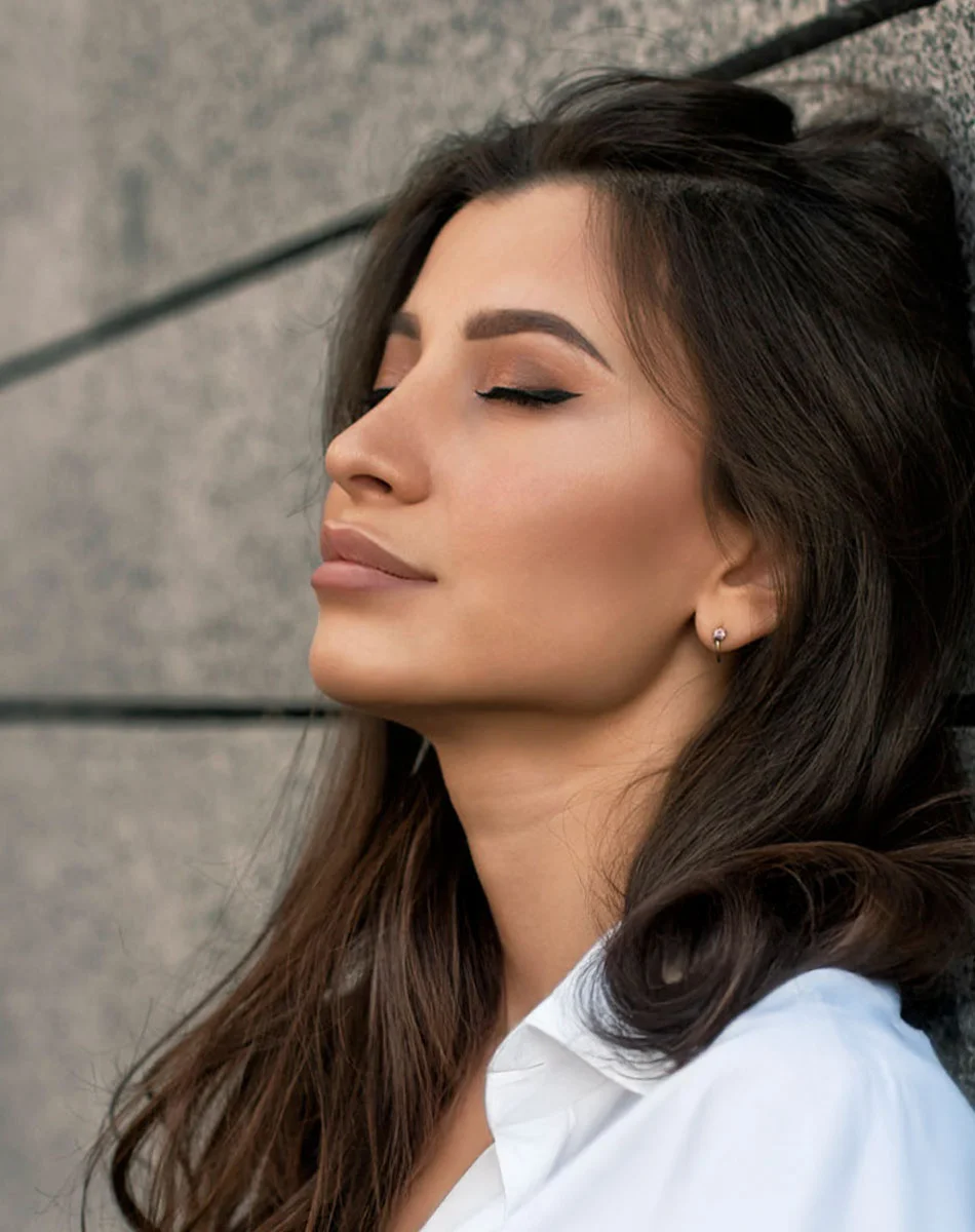 A close-up portrait of a woman with long, dark brown hair framing her shoulders and neck. Her eyes are closed and her expression is serene, suggesting a moment of relaxation or contemplation. She wears subtle makeup, including eyeliner, defined brows, and a light blush. A small stud earring is visible on her earlobe. She is dressed in a white collared shirt that rests softly at her neck. The background features a muted gray stone or tile wall, with natural outdoor lighting enhancing the peaceful mood. - Cheek Augmentation in Washington, DC and New York, NY