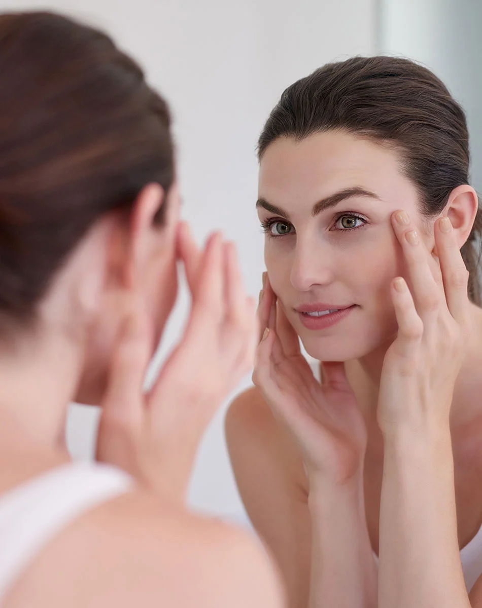 A light-skinned woman with medium-length brown hair pulled back looks at herself in a mirror with a pleasant, thoughtful expression. She wears a simple white sleeveless top and gently touches her face while focusing on her reflection. The lighting is soft and even, and the background—a neutral-toned bathroom or vanity area—is blurred, keeping the emphasis on her face and upper body. - Co2 Laser Resurfacing in Washington, DC and New York, NY