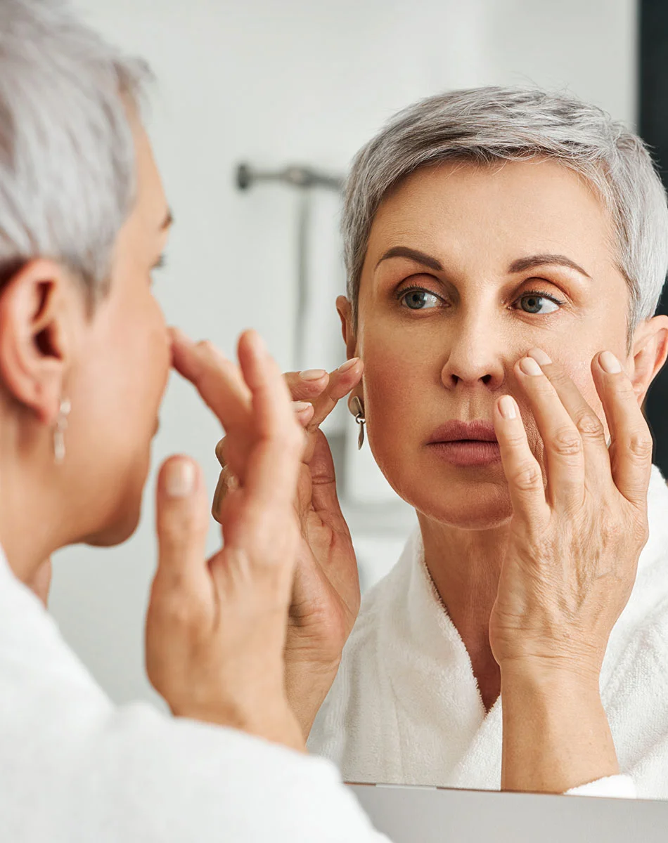 A woman with short gray hair, wearing a white bathrobe, examines her reflection in a mirror with a thoughtful or concerned expression. She gently touches the skin around her eyes and nose, focusing on its texture. Fine lines and wrinkles are visible on her face and hands, both showing natural signs of aging. The softly lit background, slightly blurred, suggests a bathroom setting. - Deeplane FaceLift in Washington, DC and New York, NY