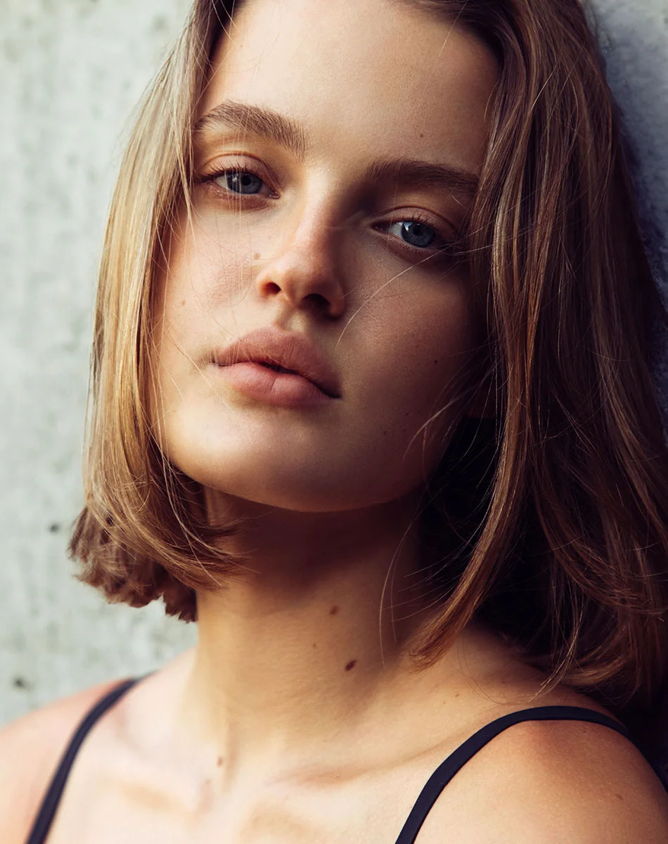 Close-up of a young woman with a short bob haircut and fair skin, wearing a black tank top. Her light blue eyes and neutral, pensive expression are framed by smooth, light-brown hair. Subtle lighting casts soft highlights on her face and neck, revealing natural features including small, light brown spots on her neck and upper chest. The background is a light gray concrete wall, adding a minimalistic, textured contrast. - Dermal Fillers in Washington, DC and New York, NY