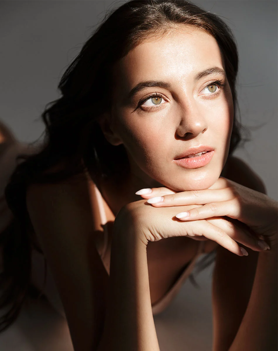A young woman with long, dark brown hair reclines against a light gray background, her face resting thoughtfully on her stacked hands. Dramatic lighting casts soft shadows across her smooth, light-toned skin, highlighting her natural glow. She wears a light peach or beige bodysuit, and her lips are a muted pink. Her nails are short with a subtle French manicure. Her gaze is directed upward and away, contributing to the introspective, elegant tone of the image. - Diamond Glow Dermal Infusion in Washington, DC and New York, NY