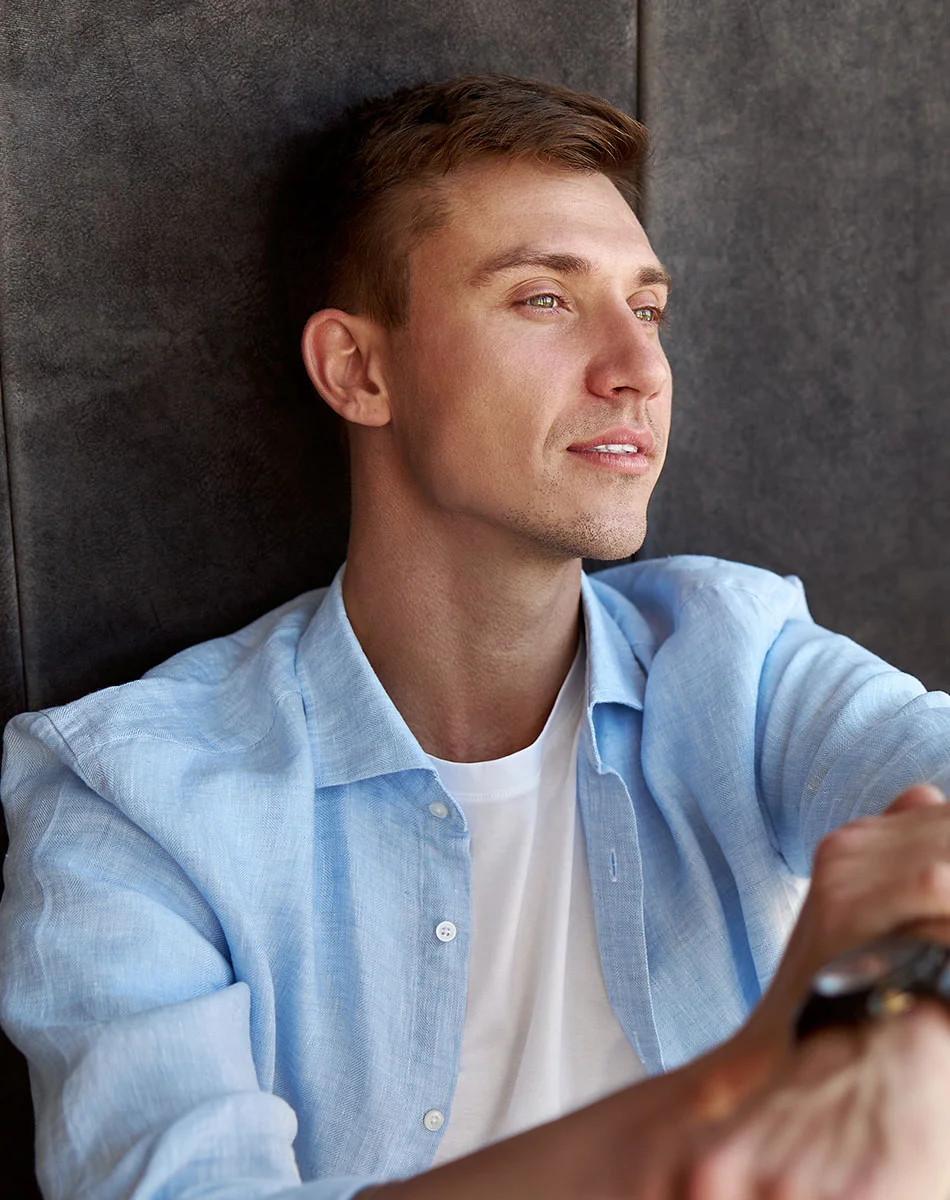 A light-skinned man with short brown hair is seen from the mid-chest up, wearing a light blue button-down shirt over a white t-shirt. He looks slightly upward to his right with a relaxed, thoughtful expression. The background is a dark gray, textured surface with natural lighting highlighting his features.- Earlobe Repair in Washington, DC and New York, NY