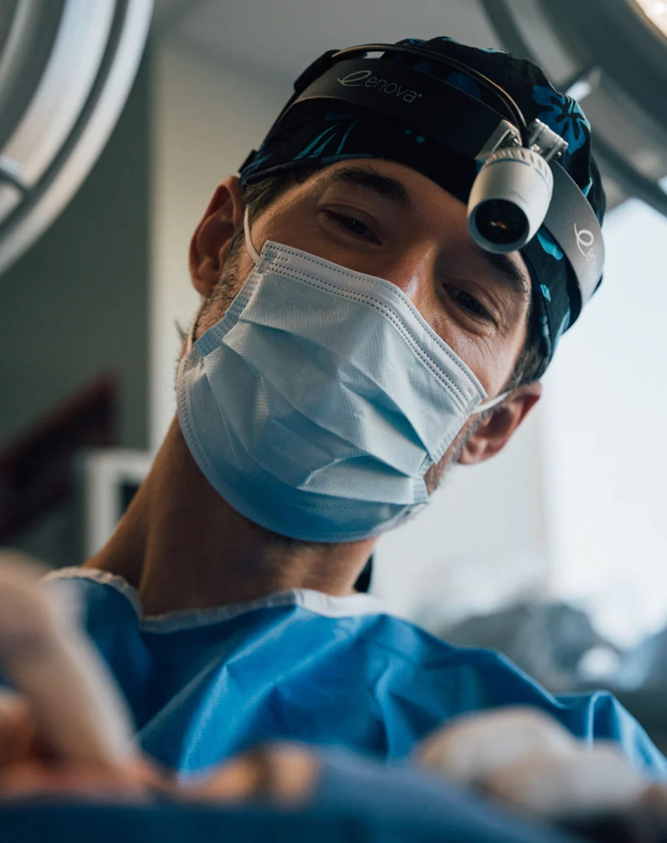 Close-up of a male surgeon wearing a light-blue gown, surgical mask, and dark surgical cap with a head lamp. He looks directly at the viewer with a pleasant expression. The background is blurred, suggesting a surgical setting. - Eyelid Surgery in Washington, DC and New York, NY