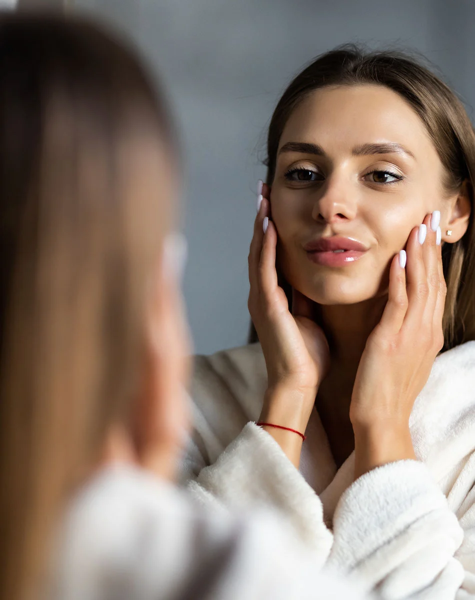 A woman with long light brown hair and a light complexion looks at herself in a mirror, wearing a cream-colored robe. Her hands gently cup her face as she inspects her skin, with a pleasant, thoughtful expression. A thin red bracelet is visible on her wrist. The background is muted gray and out of focus. - Facial Fat Grafting in Washington, DC and New York, NY
