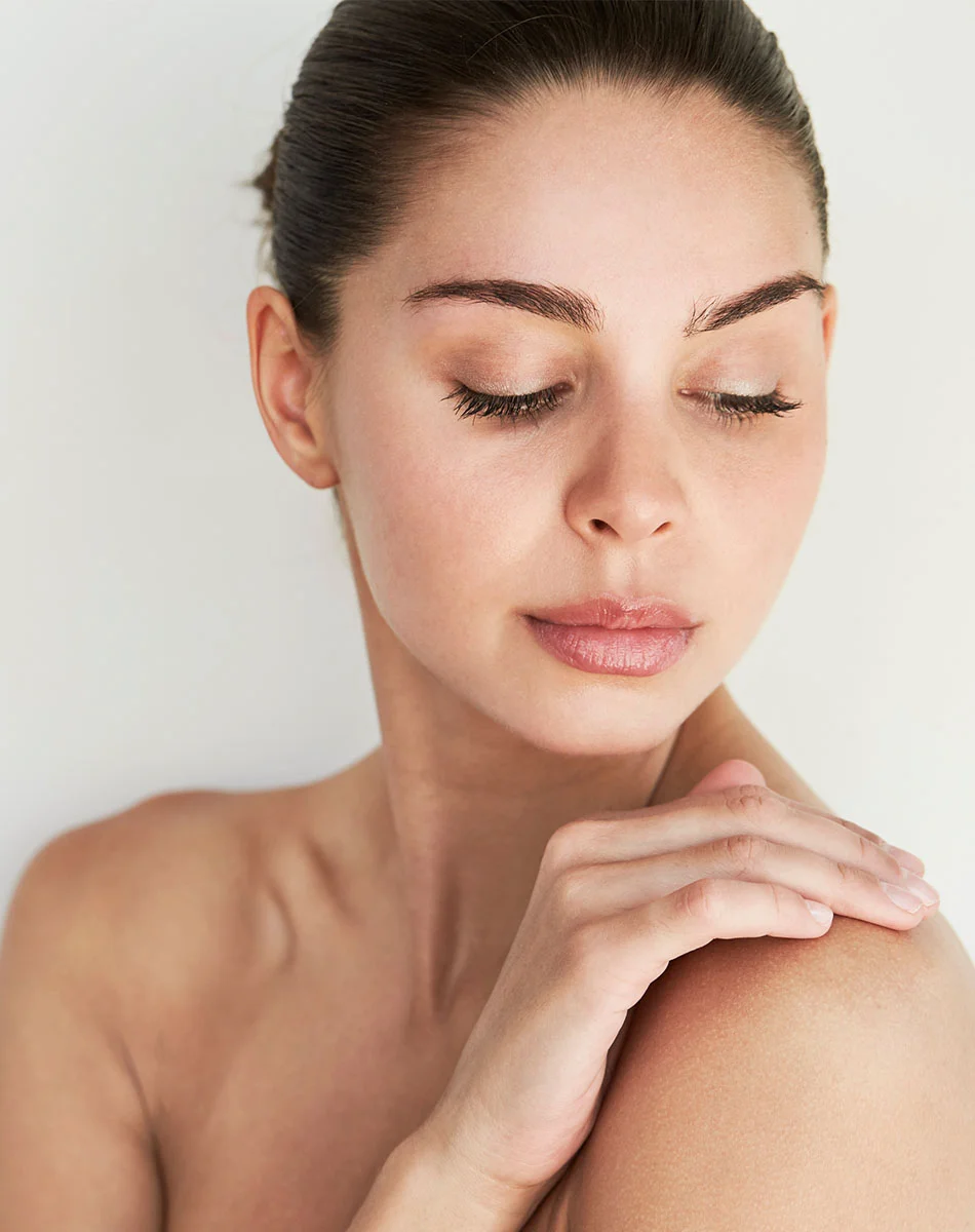 Close-up of a light-skinned woman with dark brown hair in a bun. Her eyes are closed, and she has a serene expression. One hand rests gently on her shoulder, highlighting her smooth, healthy-looking skin. The background is plain and light-colored, drawing focus to her face and upper body. - Hand Rejuvenation in Washington, DC and New York, NY