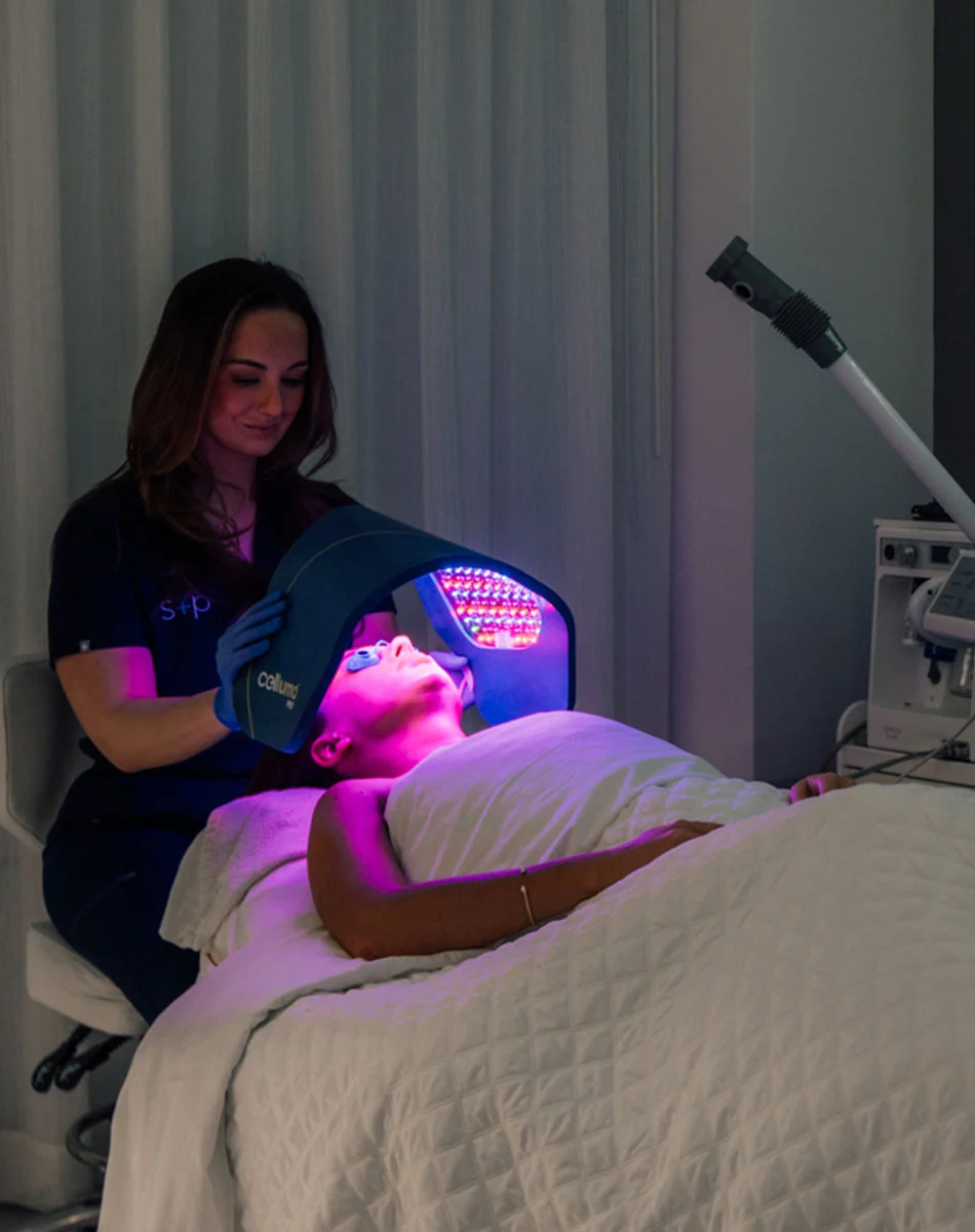 A woman lies on a light beige massage table, receiving an LED facial treatment. A professional in a dark purple/navy top holds a dark grayish-purple LED hood emitting red and purple lights over the client's face. The client is covered with a white quilted sheet, and soft pinkish-purple light glows across her face and the area. Light-colored spa equipment and vertically-lined drapes are visible in the background. - Intense Pulsed Light IPL in Washington, DC and New York, NY