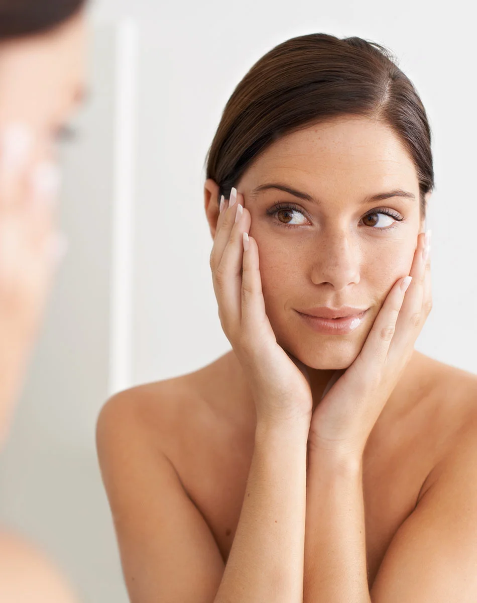 A light-skinned woman with shoulder-length brown hair is shown in a medium close-up, looking at her reflection. Her hands rest gently on her cheeks, and she has a thoughtful, slightly pleased expression. She gazes upward and to her right. Soft, natural light highlights her smooth skin and lightly colored lips. A blurred reflection is visible in the light beige background. - Laser Genesis in Washington, DC and New York, NY