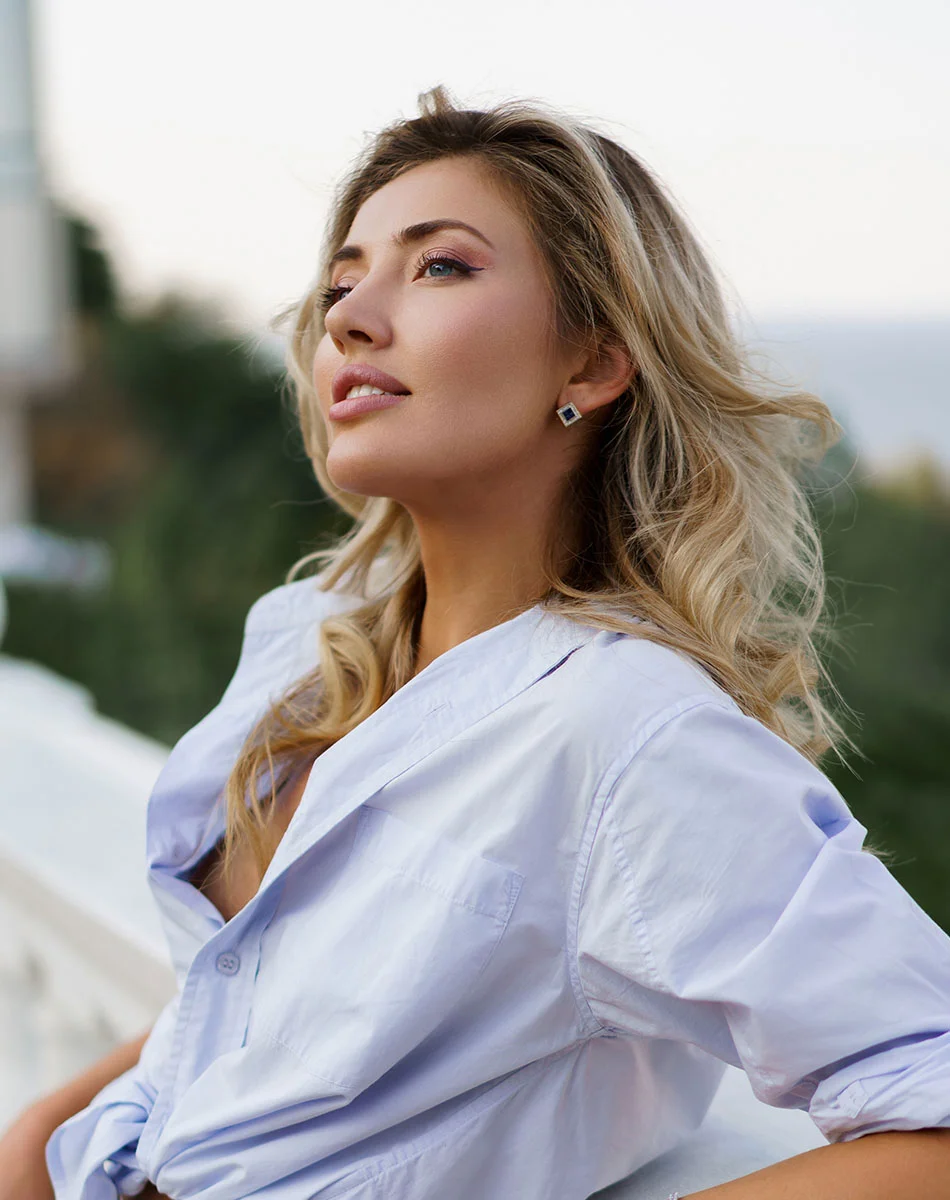 A light-skinned woman with shoulder-length blonde hair wears a light purple-blue button-down shirt tied at the waist. She has a neutral, direct expression and is looking slightly upward. Her eyes are light-colored, and she wears subtle makeup, a diamond-shaped earring, and a delicate diamond necklace. A black handbag is partially visible at her side, with a softly blurred, light-toned background suggesting an outdoor setting.- Mini Facelift in Washington, DC and New York, NY