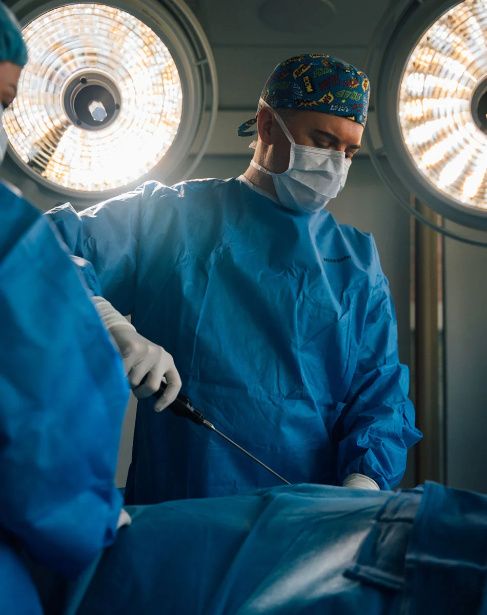 Close-up of a surgical procedure in progress. Two surgeons in blue sterile gowns and gloves are focused on the operation. The surgeon in front wears a patterned surgical cap and white mask, while another is visible in the background. A surgical instrument, possibly for a laparoscopic procedure, extends toward the draped patient. Bright overhead lights illuminate the sterile operating room. - Mommy Makeover in Washington, DC and New York, NY