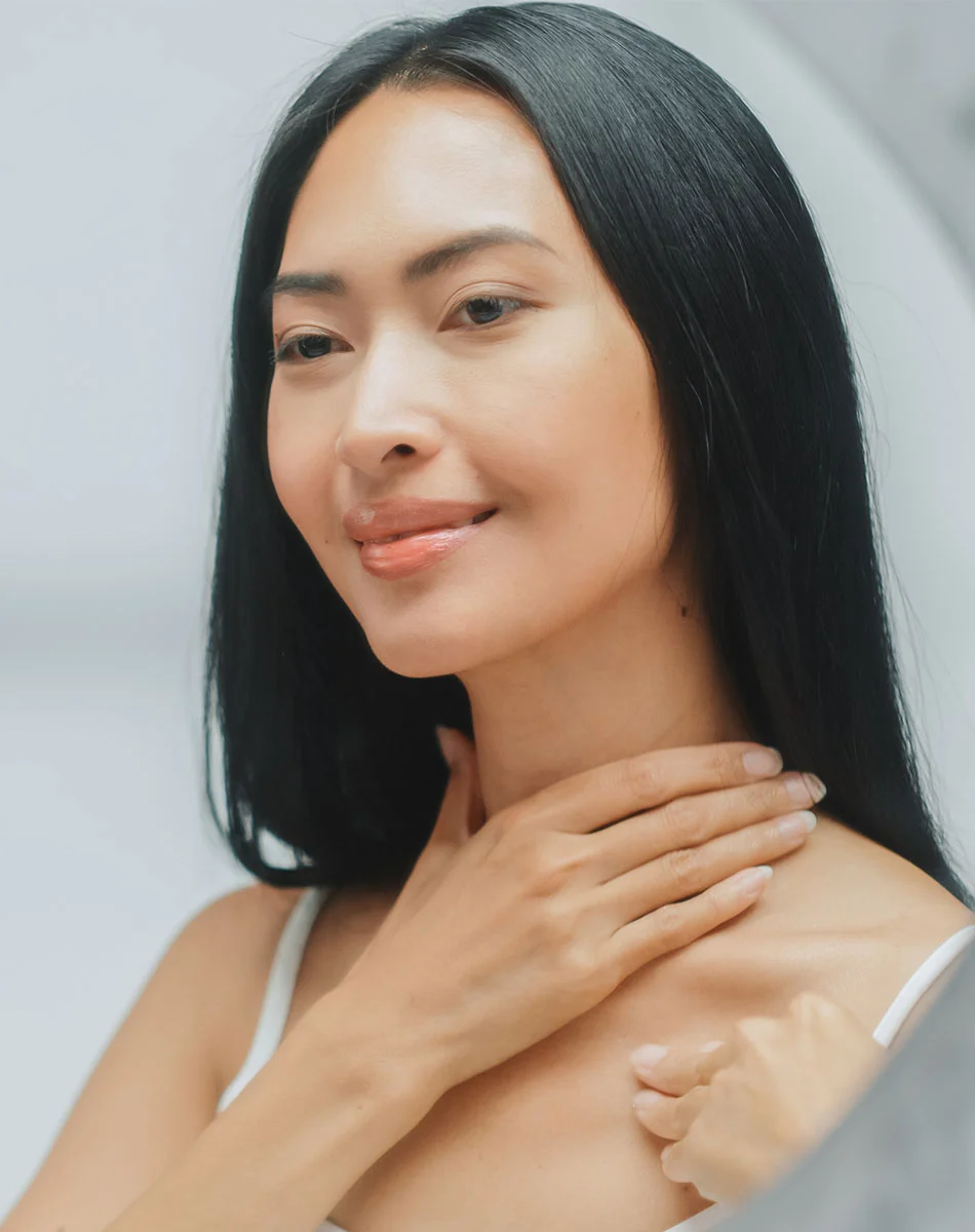 A close-up of a woman looking at her reflection. She has long, dark hair and light skin. She is smiling gently and her hands are on her neck and chest area, caressing or examining her skin. The woman appears to be examining her complexion. The background is a light, neutral color. - Necklift in Washington, DC and New York, NY