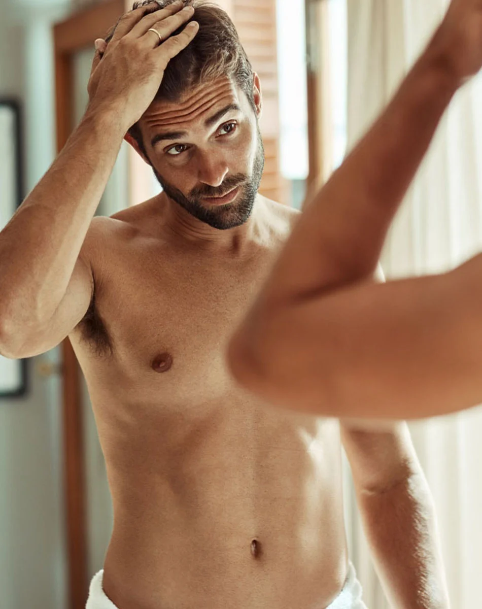 A shirtless man with short brown hair stands in front of a mirror, gently holding his hair with one hand. His expression is thoughtful, possibly concerned. He has a toned physique, and a blurred white towel is wrapped around his waist. The soft, natural lighting suggests a bathroom setting. - Non-Surgical Body Contouring in Washington, DC and New York, NY