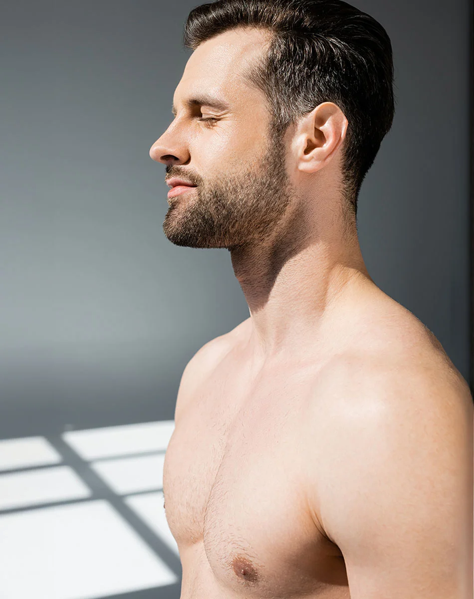 A close-up profile of a light-skinned man with short, dark brown hair and a short trimmed beard. His eyes are closed, and his expression is serene. His bare torso reveals a smooth, lightly tanned chest and upper arms. Soft, natural light creates subtle shadows on the floor, with window patterns visible in the background. The backdrop is a neutral light gray. - Pectoral Implants in Washington, DC and New York, NY