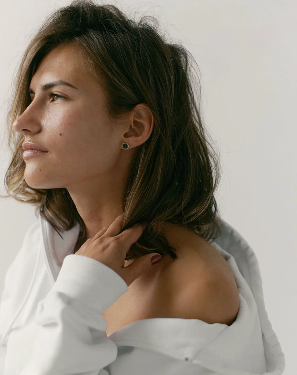 A close-up of a light-skinned woman with shoulder-length brown hair falling loosely around her shoulders. She wears a white, likely cotton, button-up shirt with slightly loose, draping sleeves. Her gaze is directed slightly to her right, and she has small, dark stud earrings. One hand rests gently on her neck and shoulder. The image is well-lit with a soft, neutral backdrop, highlighting her facial features and relaxed pose, creating a serene and aesthetically pleasing portrait. - Plasma Skin Rejuvenator in Washington, DC and New York, NY