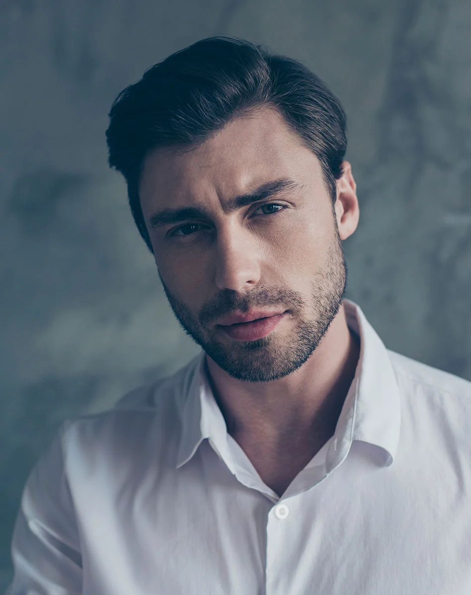 A light-skinned man with short, dark brown hair styled back is shown in a close-up portrait. His expression is neutral, almost pensive, with a slight downturn of the mouth. He gazes slightly toward the viewer and has a short dark beard and mustache. The man is wearing a button-down, white collared shirt. The background is a muted, gray-toned wall, and the lighting is even, with the focus primarily on his face and upper torso. - PRP Hair Maintenance in Washington, DC and New York, NY