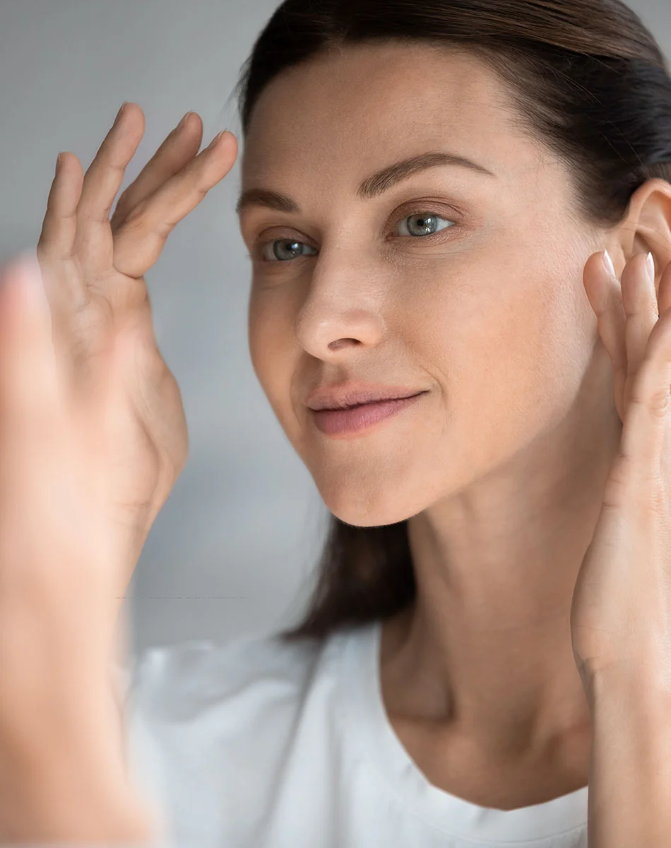 Close-up of a woman examining her face in a mirror. The woman has light brown hair pulled back.  She is looking at herself in the mirror, her hands are near her face, one hand is on her forehead and the other near her ear. The background is a light, neutral gray.- Revision Facelift in Washington, DC and New York, NY