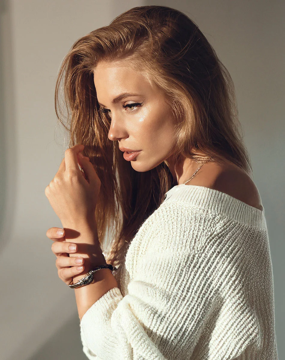 A woman with light brown, shoulder-length hair wears a cream knit sweater and bracelet. She looks slightly left with a neutral expression. Soft natural light and a pale gray background complete the scene. - Revision Rhinoplasty in Washington, DC and New York, NY