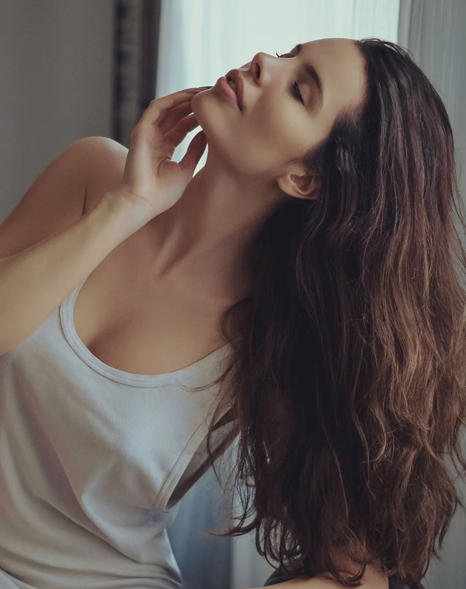 A young woman with long, dark brown wavy hair is seen from the mid-chest up. She is wearing a light gray or white tank top. Her eyes are closed, and her head is tilted back slightly. Her hands are gently resting on her neck and jawline. The lighting in the image is soft and natural, with a window visible in the background. A soft focus effect is also present. The woman's expression suggests a peaceful or relaxed state. - Sculptra in Washington, DC and New York, NY