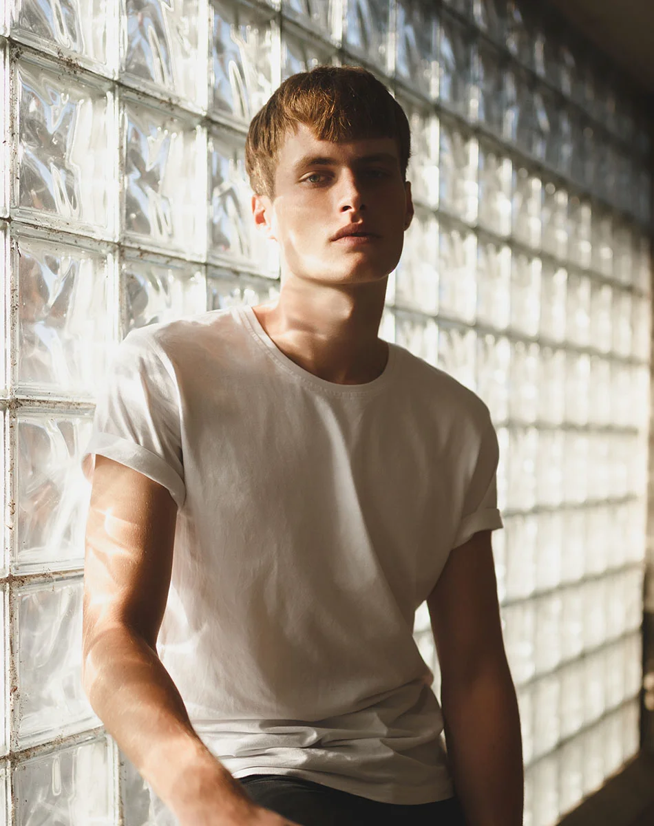 A young man with short light brown hair leans against a wall of large, translucent glass blocks. He wears a plain white short-sleeved t-shirt and looks directly at the camera with a neutral expression. Sunlight filters through the glass, casting soft highlights and shadows on his skin and shirt, with faint sun-kissed patterns visible on his arms and shoulders. - Teenage Rhinoplasty in Washington, DC and New York, NY
