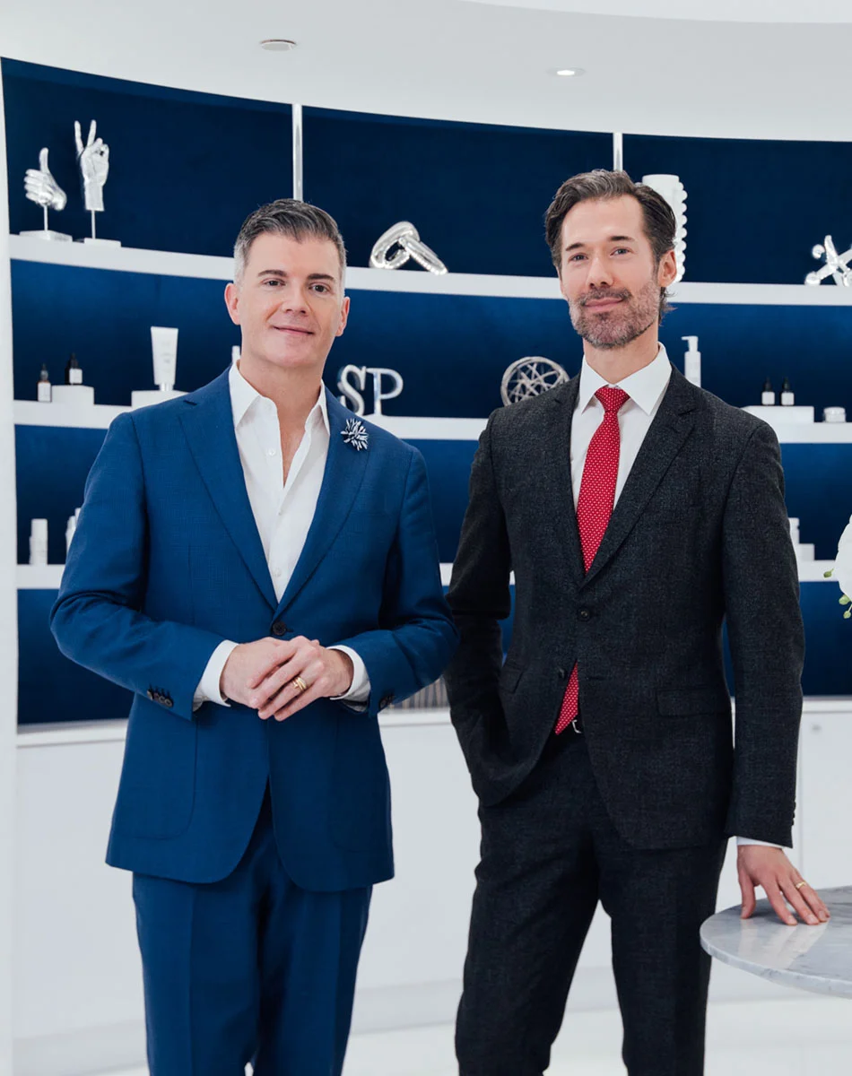 Dr. Somenek and Dr. Pitman stand in a bright, modern interior. Dr. Somenek wears a deep blue suit with a white shirt and a decorative pin, while Dr. Pitman wears a dark suit with a red tie. Behind them are navy shelves with sculptural decor. The space has light-colored walls and flooring, suggesting an office or medical setting. - OOT Recovery in Washington, DC and New York, NY